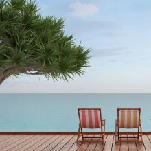 image of two striped lounge chairs on a wooden deck overlooking a calm ocean beside a palm tree via Canva