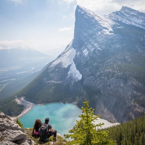 two people sitting side by side on the edge of a rocky cliff overlooking a lake and snowcapped mountains Photo by Kalen Emsley on Unsplash