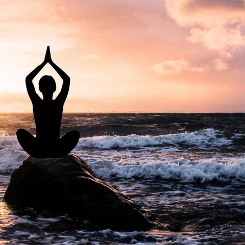 a person holding hands overhead in prayer pose while seated on a rock beside the ocean at sunset