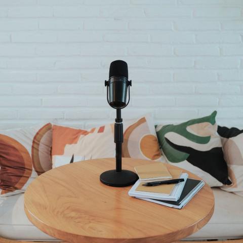 a standup microphone on a small round wooden table with books and a pen in front of a bench seat with colorful pillows via Canva