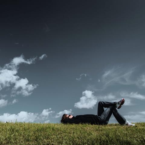 photo of a person lying on their back on grass with one leg crossed over the other knee looking up at a partly cloudy sky via Canva