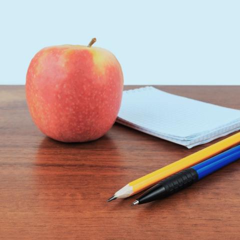 a pen and pencil beside a small notebook and a red apple on a wooden surface via Canva