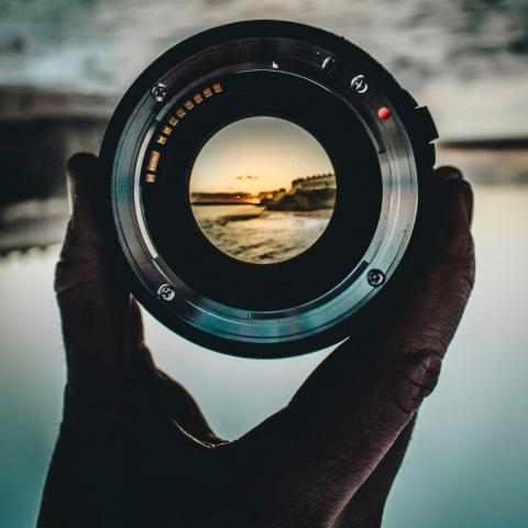 silhouette of a person's hand holding a camera lens to bring distant sunset over water into focus via canva