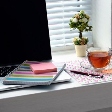 desk with open laptop, multicolor striped notebook, pink sticky notes, pen, cup of tea in glass mug, potted plant, and light filtering through white blinds, via Canva