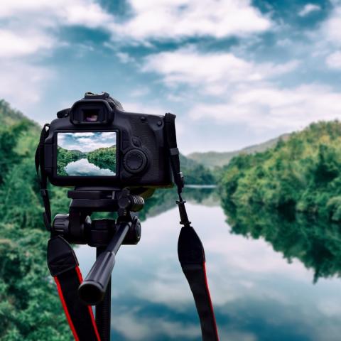 photo of a camera on tripod aimed at a river flanked by trees under partly cloudy sky via Canva