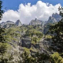 September 2021 photo writing prompt contest image_tall jagged mountains and waterfalls against blue sky and behind trees via Photo by Marius Girard on Unsplash