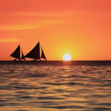 two sailboat silhouettes against a red sky at sunset with the sun just dipping below the horizon of the ocean 