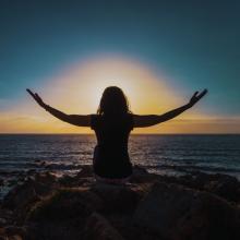 Silhouette of a seated person with arms outstretched before the ocean at sunset