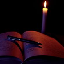 dark photo of a single pillar candle behind an open book with a pen resting on it, via Canva