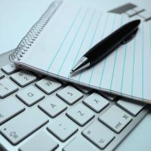 photo of a Mac keyboard with a lined spiral bound notebook and black pen lying on top via Canva