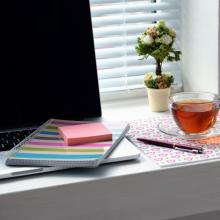 desk with open laptop, multicolor striped notebook, pink sticky notes, pen, cup of tea in glass mug, potted plant, and light filtering through white blinds, via Canva