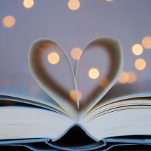 an open book with pages folded in to resemble a heart and twinkling lights in the background via canva